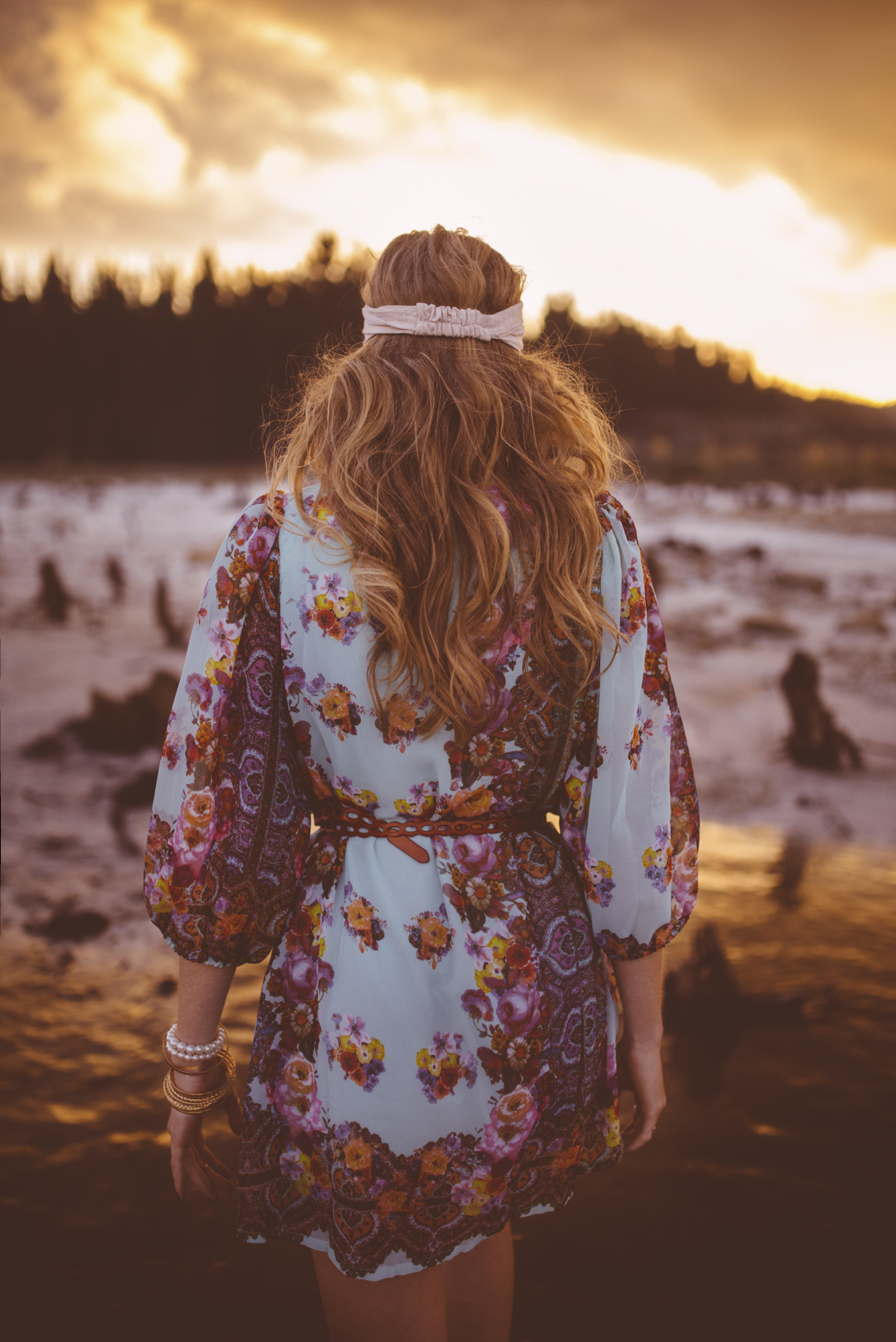 Rearview of a boho girl wearing a floral dress walking through water in nature at sunset
