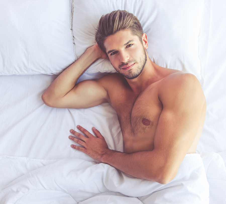 Top view of sexy muscular young man looking at camera with sensual smile while lying in bed